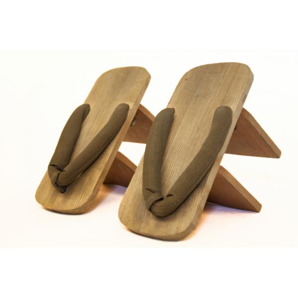 A pair of stilted thong shoes on an angle. The wood is light brown and the fabric for the thong is brown. Two stilts are visible on the bottom of each shoe. 