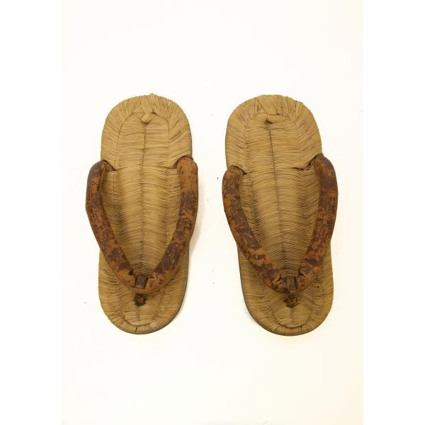 A pair of light brown flat thong sandals. Rice straw has been woven together to create the bed of the shoes and a piece of brown material forms the thong. It is attached to the front in the centre and two-thirds of the way back to both sides. 