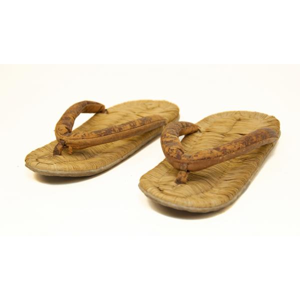 A pair of light brown flat thong sandals. Rice straw has been woven together to create the bed of the shoes and a piece of brown material forms the thong. It is attached to the front in the centre and two-thirds of the way back to both sides. 