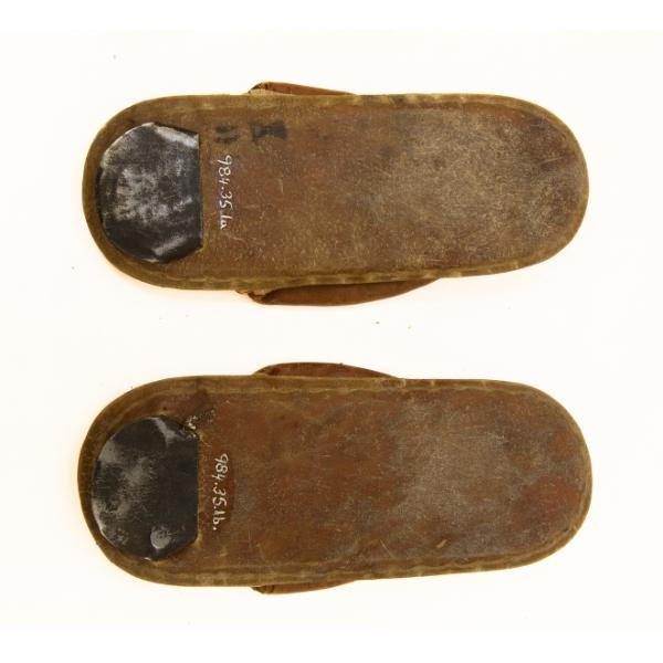 The bottom of pair of flat thong sandals. The sole is brown and has been sewn to the upper part. There is a piece of black material towards the toe of each shoe