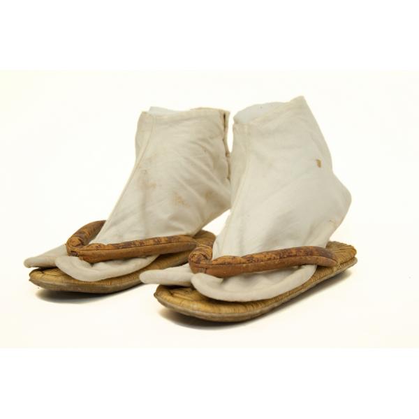 A pair of white cotton socks inside a pair of brown flat sandals. The toe compartment is split into two sections, with one section on either side of the thong. 