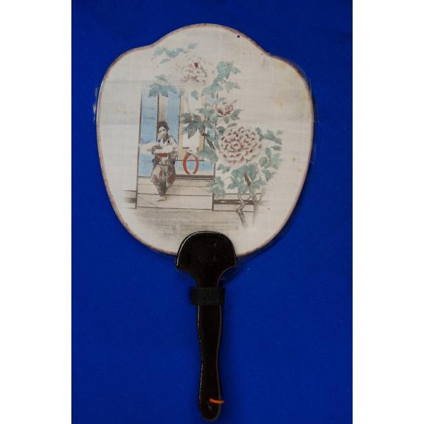 A white fan with a black handle spread open on a blue background. An image of a Japanese woman inside the door of a building with a flowering tree outside is printed on the fan. 