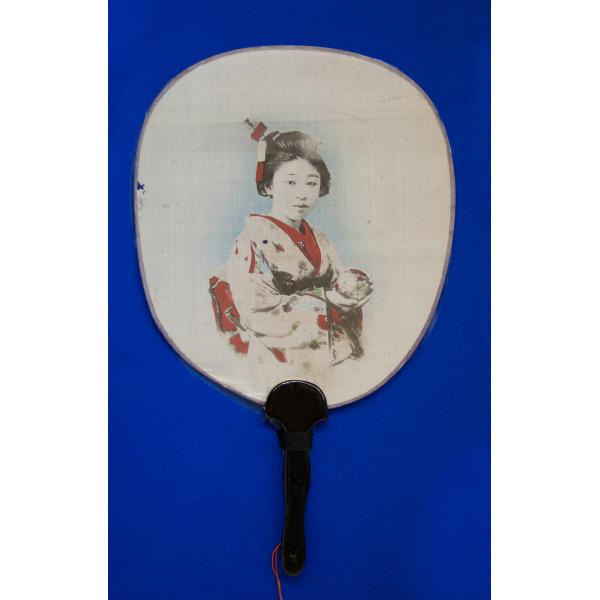 A white fan with a black handle spread open on a blue background. There is an image of a Japanese woman in traditional clothing, from the waist up, printed on the fan. 