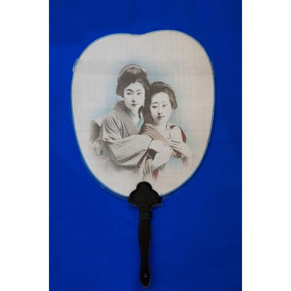 A white fan with a black handle on a blue background. The image of two Japanese women in traditional clothing, from the waist up, is printed on the fan.  