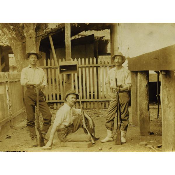 Three men in light shirts and dark pants, holding guns, in front of a wooden picket fence. The man in the centre is sitting on the ground and the other two are standing. 