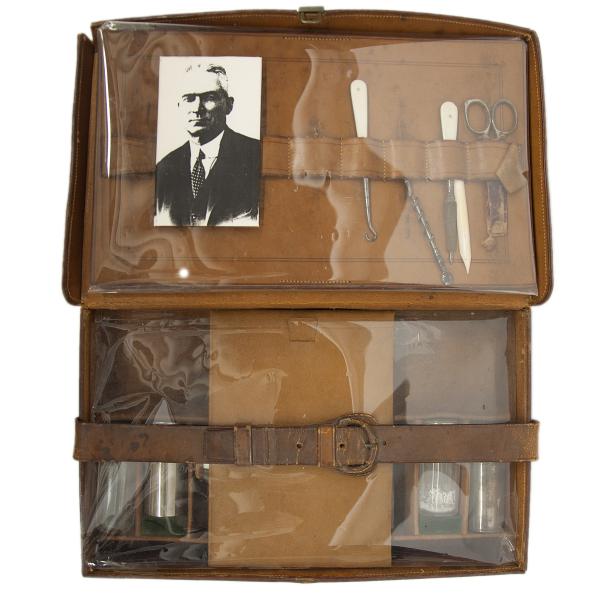 Looking at a leather personal travel case from above. There is a belt across the bottom to secure the contents. The top portion has a photo of a main in a suit as well as scissors and other small tools.