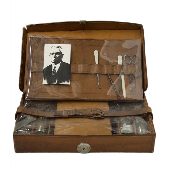 An open leather case with a metal latch on the front and removable organizers. There is a photo of a man in a suit along with scissors and other tools in the top part of the case. There is a belt across the bottom section. 