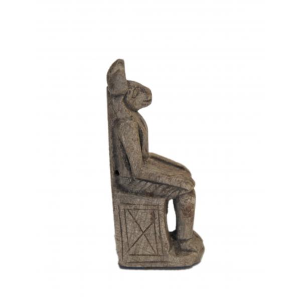 Vue de côté d’une statuette d’Anubis assis. Il a de grandes oreilles sur le dessus de la tête et son visage est visible de profil. Le bas de ses jambes est éloigné du trône, créant un espace. 