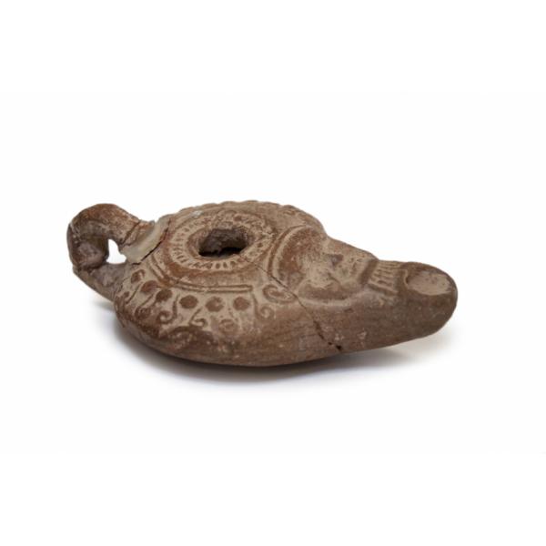 Side view of the terracotta oil lamp. There is a handle at one end and a spout at the other that has a face on it. There is an opening in the middle of the lamp surrounded by detailing. 