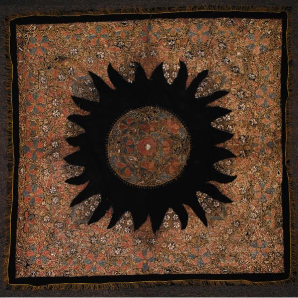 A tablecloth embroidered with gold, pink, green, and white thread. There is a black sunburst around a medallion in the centre. Outside the sunburst there is a large square of embroidery, followed by a black border and an embroidered border.