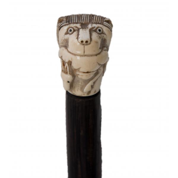 Close-up view of an ivory handle carved into a lion's head with a small horse underneath. It is attached to a dark wooden cane.