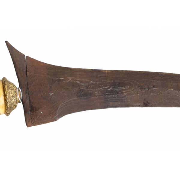 The metal blade of a dagger and the brass-coloured piece connecting it to an ivory handle. The blade is rusty and has there is a swirling pattern in the metal.  