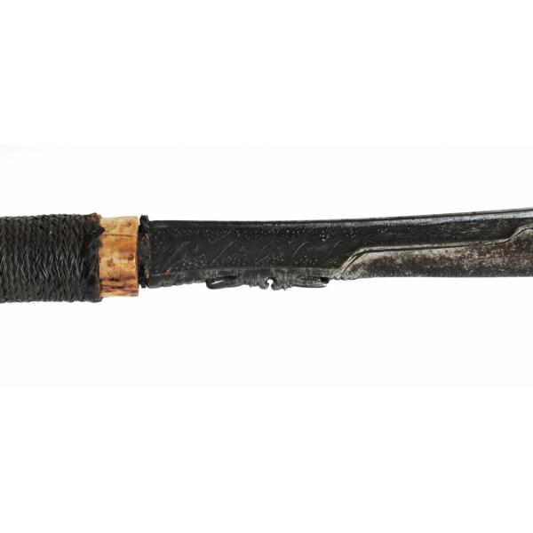 The blade of an axe. It has a rattan braid on the handle above it. The blade has a pattern of stamped scroll-work on it.