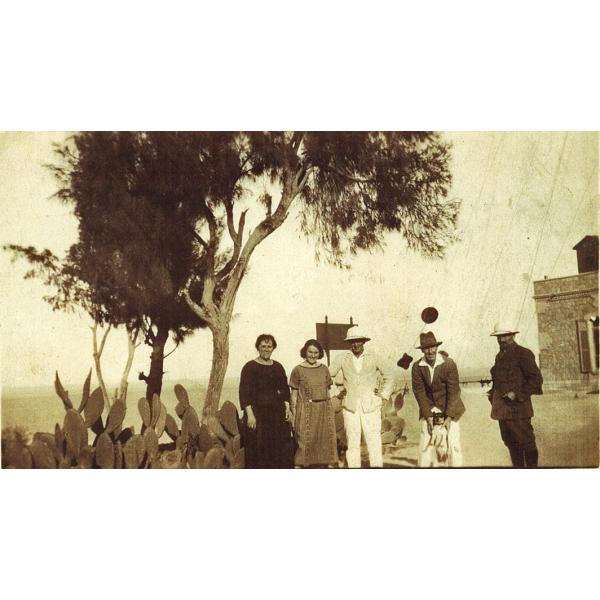 Two women in long dresses standing beside three men in suits and hats. There is a tree behind them and the corner of a stone building is visible.