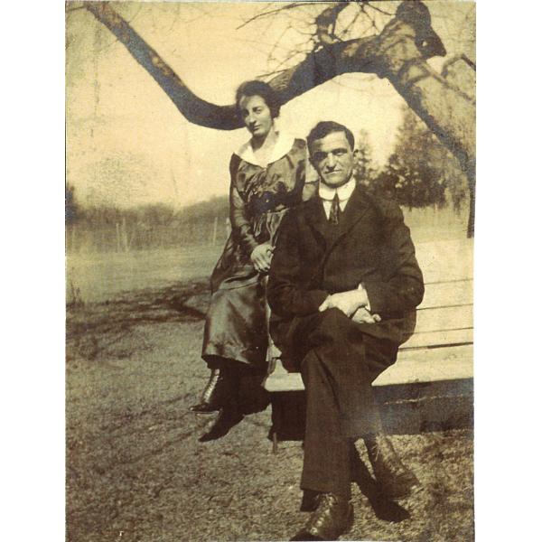 A woman in a long, dark dress and a man in a dark suit and tie sit on wooden planks beneath a tree branch.