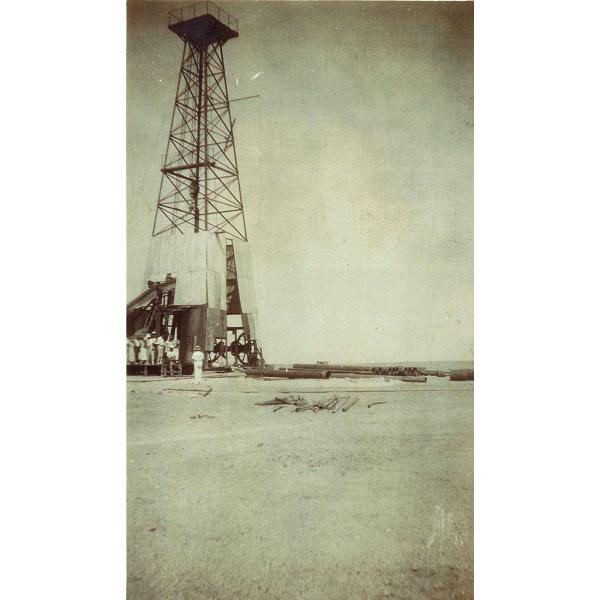 A group of people stand at the base of an oil rig with a platform at the top. The bottom part of the rig is covered with sheets of metal. There is a pile of casing off to the right side.