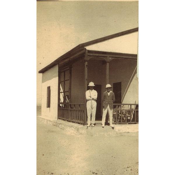 Une photo de deux foreurs internationaux debout à côté d'une maison blanche avec du bois foncé le long des bords et de la balustrade du porche. Ils portent des casques coloniaux blancs. L'un porte une chemise blanche et l'autre une veste foncée. 