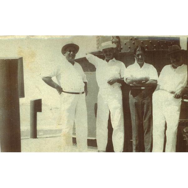 A photo of four men dressed in white on the deck of a ship. They are standing in front of a metal column with rivets in it.  