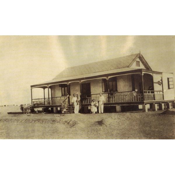 Two men, a woman, and two children standing in front of a small house with a porch. They are all wearing white. There is sand in the foreground and background. 