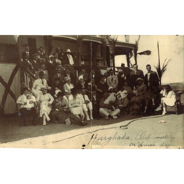 A group of people posed in the shade of a building, but the steps. The men are in suits and the women are in dresses.