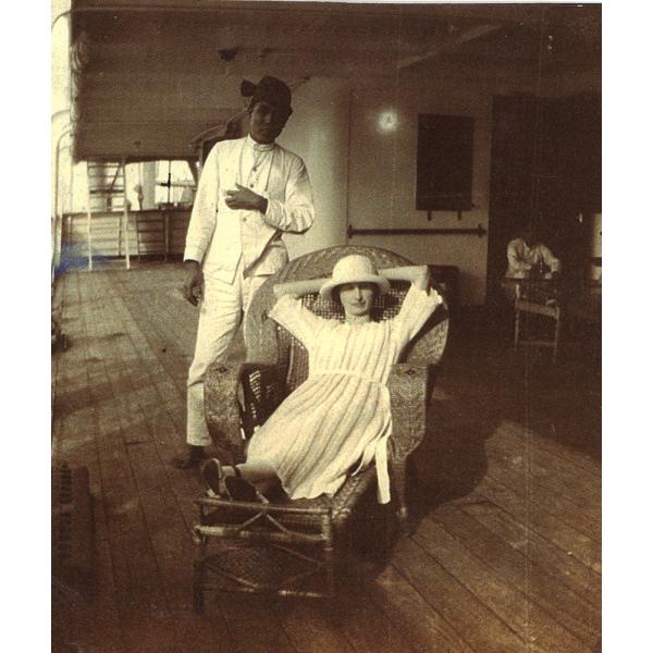 Une femme allongée dans un fauteuil inclinable en osier, portant une robe blanche et un chapeau. Un homme vêtu d’un costume blanc se tient derrière elle. Ils sont sur le pont d’un bateau au plancher de bois.