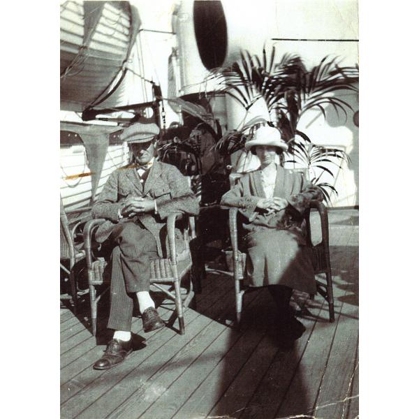 A man and a woman sitting on the deck of a ship. There are life boats hanging to the left and a palm frond behind them. The floor is made from wooden boards.