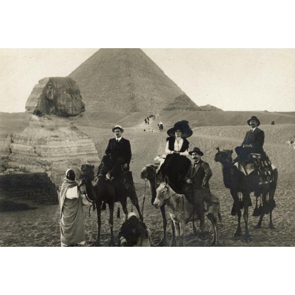 A postcard of four people riding three camels and a donkey in front of the Sphinx and a pyramid. 