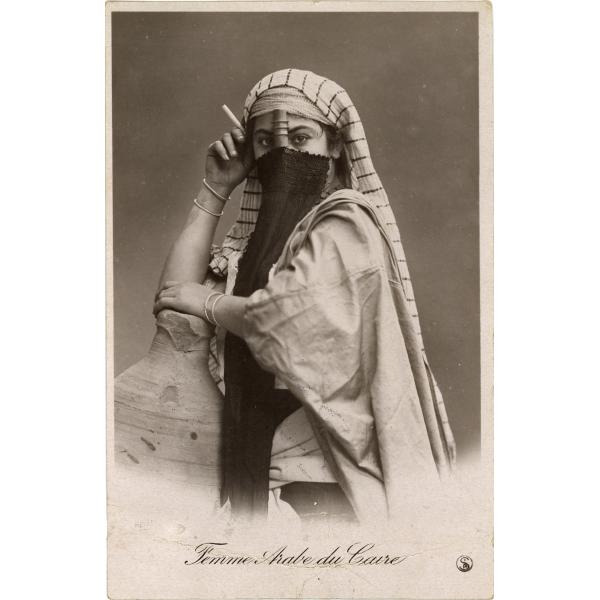 Une carte postale avec une femme voilée portant une étoffe rayée blanche sur la tête. Elle porte deux bracelets, un sur chaque poignet, et tient une cigarette. L’inscription au bas indique : « Femme arabe du Caire »  