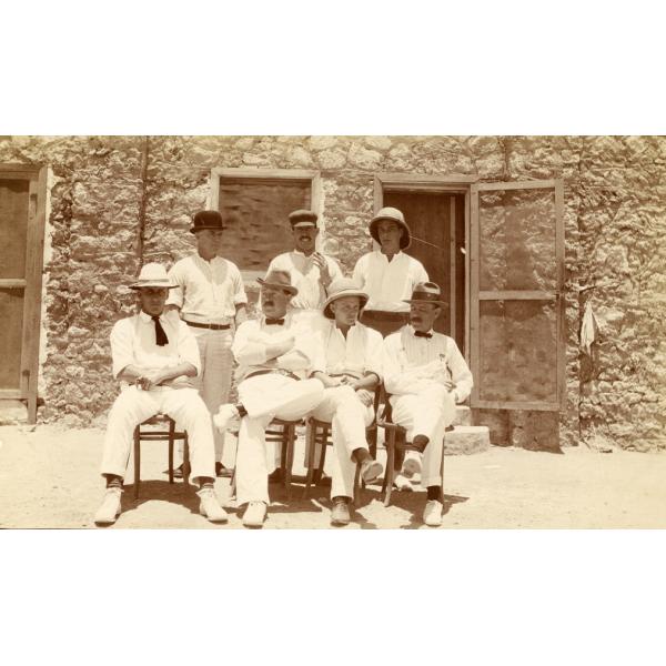 A photo of three men standing and four men sitting in front of a stone building with two screen doors and a window. They are wearing white and various types of hats.