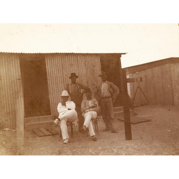 Four men in front of a small building. Campbell and Gillespie are seated, while Gardiner and Kerby are standing. They are wearing an assortment of work clothes.