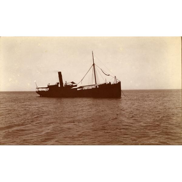 A photo of a ship on camp water. It is listing slightly to the side and the anchor is down to the right. There is one mast and a smokestack.