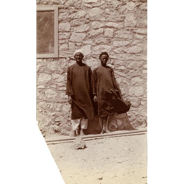 Une photo de deux hommes portant des robes foncées, debout devant un mur de briques avec une fenêtre munie d’une moustiquaire.  