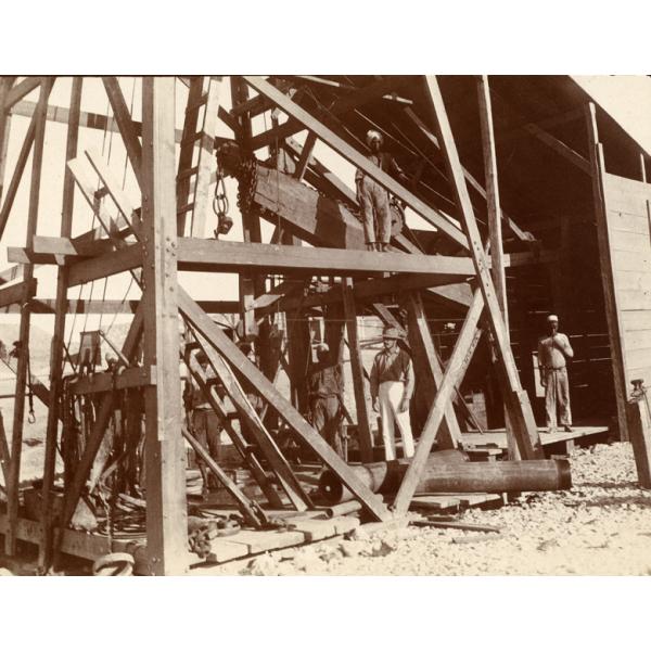 William Gillespie and his workers underneath a wooded rig on Jubal Island. Gillespie is wearing white pants and a pith helmet. 