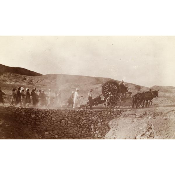 Une photo de deux chevaux tirant une charrette munie de deux grandes roues. Un groupe d’hommes se tiennent à l’arrière et tirent une corde. Une route a été construite avec de petites roches pour traverser un fossé.  