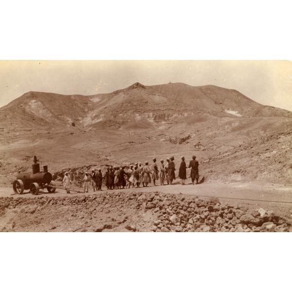 A photo of Egyptian workers standing in a line, hauling a boiler with ropes along a road in the desert. The boiler has four weeks and is has two cylindrical components put together. 