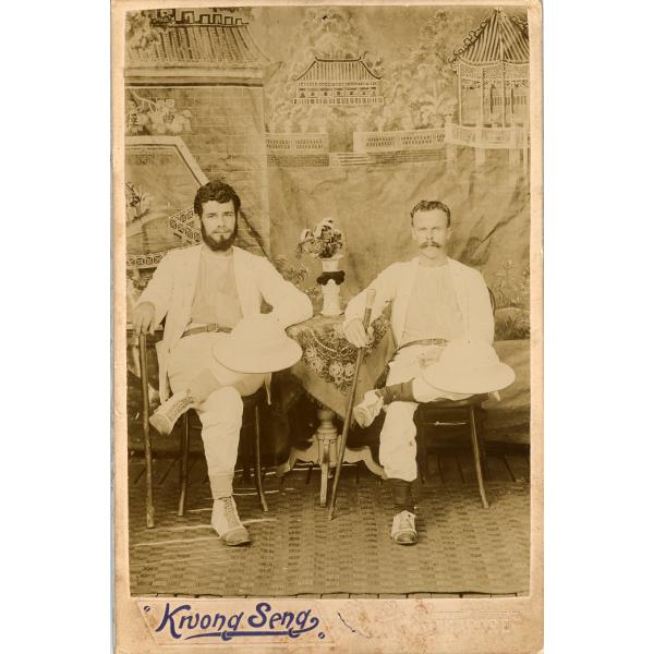 A photo of two men wearing white, sitting cross-legged on chairs on either side of a small table covered with a cloth. There is a vase of flowers on the table. Both men have a cane in their right hand and are balancing pith helmets on their left knee. There is a painted background of temples in the background.