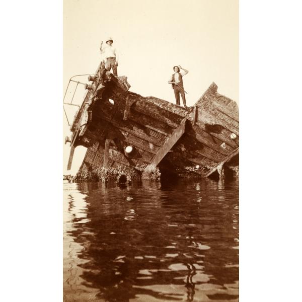 A photo of two men standing on top of a ship wreck in the water. Only part of the hull is visible.