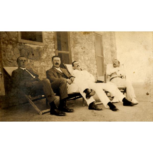 A photo of four men sitting in reclining chairs in front of a stone building. The two on the left are wearing dark jackets and the two on the right are in white.