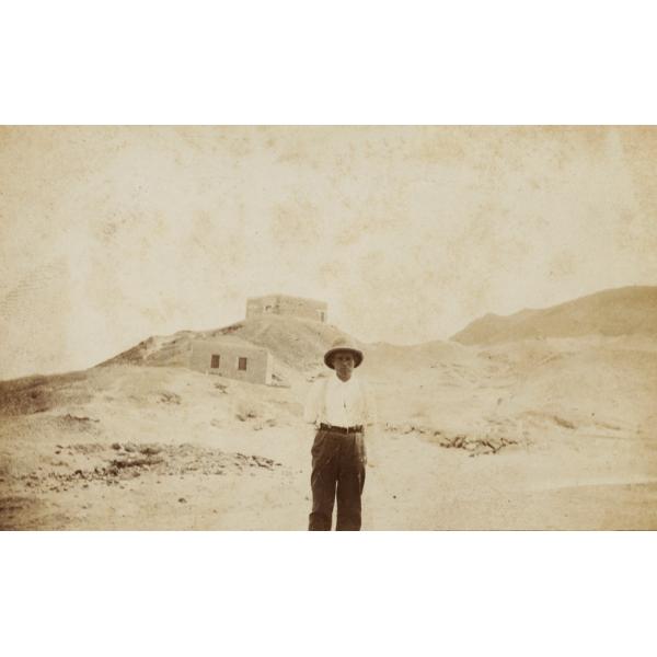 A photo of an International Driller standing in the desert with his arms at his sides. He is wearing a white shirt, dark pants, and a pith helmet. There are two small buildings on a hill in the background.  