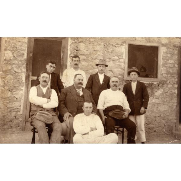 A photo of eight men in front of a stone building with a wooden-framed door and window. Four are standing at the back, three are sitting in chairs, and one is sitting on the ground.