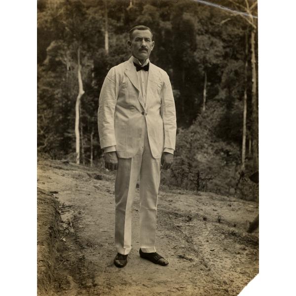 A photo of William O. Gillespie in a white suit with a dark bow tie and shoes. He is standing on a path in front of trees. 