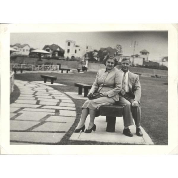 A photo of Vola and John Braybrook sitting on a park bench. There is a path with large stones beside them and a series of benches along the path behind them. She is wearing a skirt and jacket with high heels and he is wearing a suit and tie.