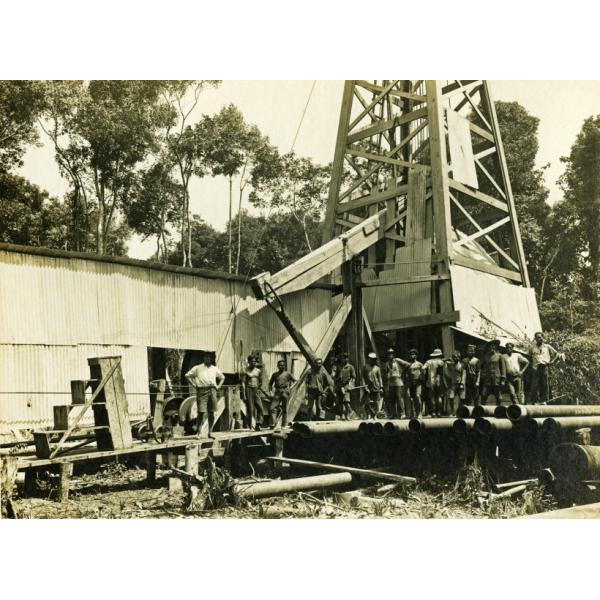 A group of men standing in front of an oil rig that is covered in sheet metal. There is a pile of casing in front of them and there are trees in the background. 
