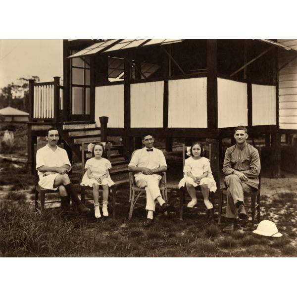 Three men and two girls (alternating) sit in chairs outside a building on stilts. They are wearing light clothing. The building has a porch with stairs and white and dark railings and posts.  