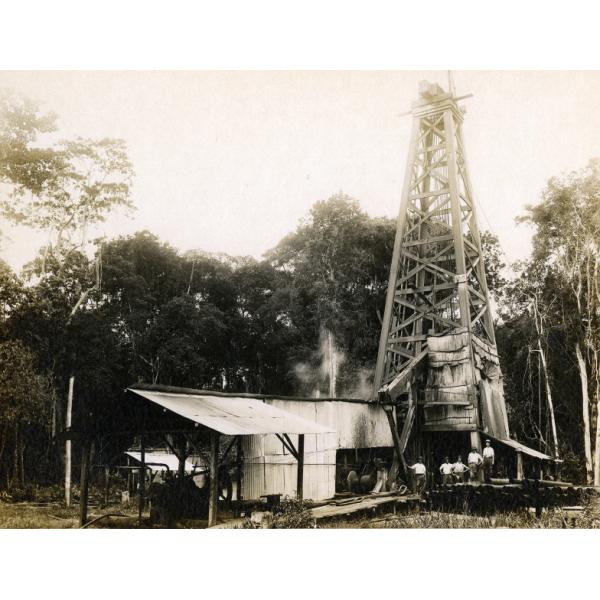A photo of five men standing by a pile of casing in front of a drilling rig. There is a structure with a roof in the foreground, and a line of trees in the background.