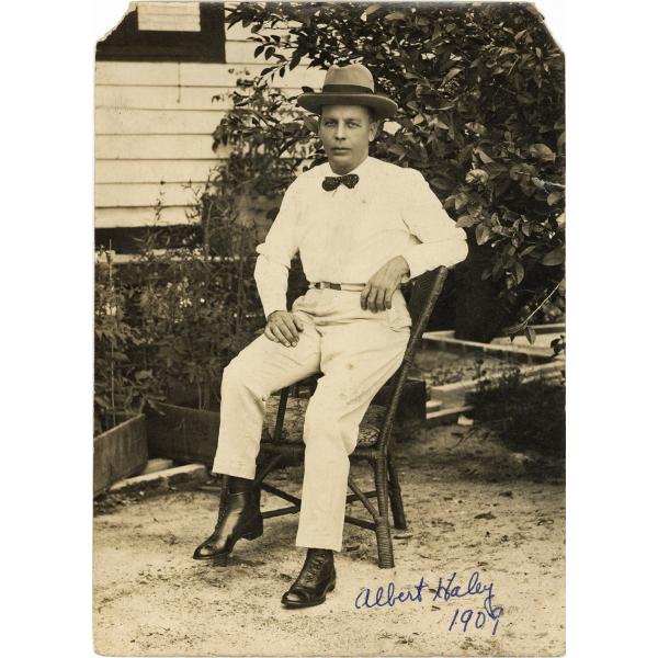 A photo of Albert Haley sitting on the arm of a chair. He is wearing white pants, a white shirt, a bow tie, and a hat. There are bushes behind him as well as a building with white siding.