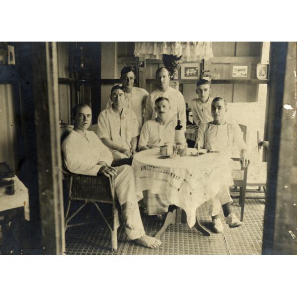 A photo of seven International Drillers around a table indoors with a white tablecloth. They are wearing white and four are sitting on wicker chairs. There is a cup, a pipe, and a bottle on the table.