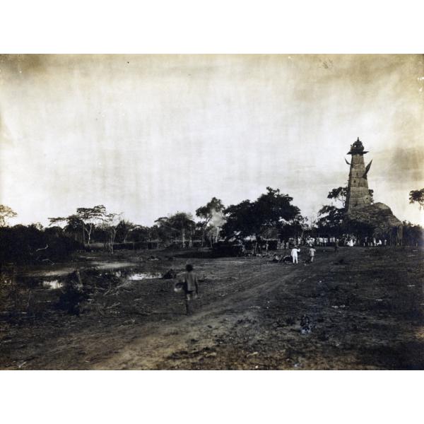 A photo of a boy walking down a road towards an oil derrick covered in thatching. There is a large crowd underneath the derrick. The area beside the road is empty and there are trees in the background.