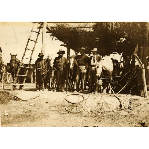 A photo of four men who are wearing hats standing with five horses, underneath a structure with a roof made from vegetation that has a wooden ladder leaning against it.