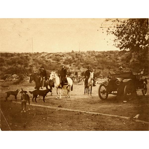 Three men on horses. There are three dogs in front of them and a long pipe that is half-buried. A fourth man sits in a car to the right. There is a hill with bushes on it in the background.